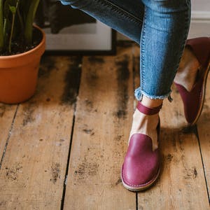 Op de afbeelding: Een paar bordeauxrode leren enkelriem sandalen met een gesloten neus. De sandalen worden gedragen door een persoon met blote benen en blauwe jeans.