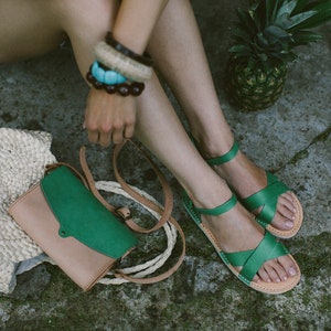 May include: A pair of green leather sandals with a woven strap design. The sandals are worn by a person sitting on the ground with a straw bag and a green and brown leather bag.