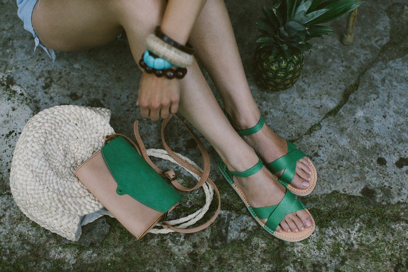 May include: A pair of green leather sandals with a woven strap design. The sandals are worn by a person sitting on the ground with a straw bag and a green and brown leather bag.