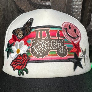 Op de afbeelding: Witte en zwarte truckerpet met geborduurde patches. De patches bevatten een vlinder, bloemen, sterren en een smiley-gezicht. De belangrijkste patch toont een tractor en de woorden "Farm Girl" in een decoratief lettertype.