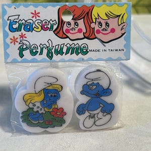 May include: Vintage "Eraser Perfume" package featuring two Smurf erasers. One eraser depicts Smurfette with a flower, and the other shows a male Smurf. The package has cartoon illustrations and text that reads "Made in Taiwan."