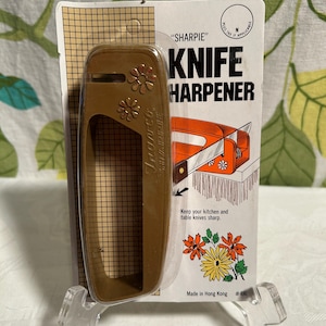 Pode incluir: Um afia-facas de pl&aacute;stico castanho com um design floral. A embalagem diz "SHARPIE" KNIFE HARPENER. Diz "Keep your kitchen and table knives sharp." e "Made in Hong Kong #490".
