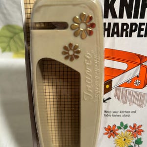 May include: A beige plastic knife sharpener with a gold flower design. The packaging says "SHARPIE" KNIF HARPER and "Keep your kitchen and table knives sharp." It also says "Made in Hong Kong #490".