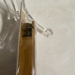 Pode incluir: Close-up de uma caixa dourada com uma tampa de pl&aacute;stico transparente. A caixa tem um mecanismo de mola vis&iacute;vel no lado.