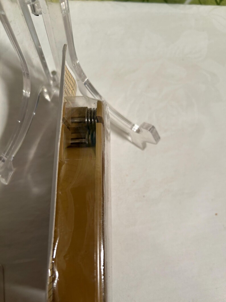 Pode incluir: Close-up de uma caixa dourada com uma tampa de pl&aacute;stico transparente. A caixa tem um mecanismo de mola vis&iacute;vel no lado.