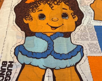 Op de afbeelding: Een stoffen patroon voor een pop met bruin haar, een blauw-witte jurk en oranje schoenen. Het patroon bevat de tekst "Bubbles" en "Hugga Bunca".