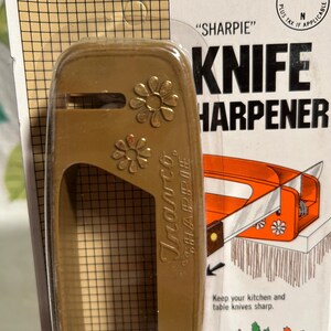 Pode incluir: Um afia-facas de pl&aacute;stico castanho com um design floral. O afia-facas est&aacute; embalado num cart&atilde;o com o texto "SHARPIE" KNIFE HARPENER. O cart&atilde;o tamb&eacute;m inclui instru&ccedil;&otilde;es para manter as suas facas de cozinha e de mesa afiadas.