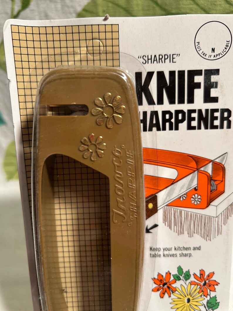 Pode incluir: Um afia-facas de pl&aacute;stico castanho com um design floral. O afia-facas est&aacute; embalado num cart&atilde;o com o texto "SHARPIE" KNIFE HARPENER. O cart&atilde;o tamb&eacute;m inclui instru&ccedil;&otilde;es para manter as suas facas de cozinha e de mesa afiadas.