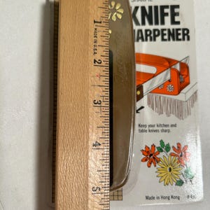 Pode incluir: Um afia-facas de pl&aacute;stico castanho com um design floral e o texto "SHARPIE" KNIFE SHARPENER. O afia-facas est&aacute; embalado em uma embalagem blister de pl&aacute;stico transparente com fundo branco e o texto "Made in Hong Kong #491".