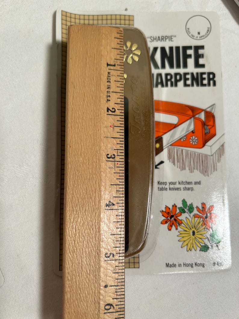 Pode incluir: Um afia-facas de pl&aacute;stico castanho com um design floral e o texto "SHARPIE" KNIFE SHARPENER. O afia-facas est&aacute; embalado em uma embalagem blister de pl&aacute;stico transparente com fundo branco e o texto "Made in Hong Kong #491".
