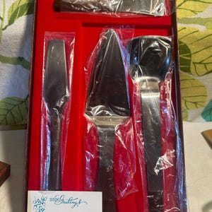 May include: A four-piece stainless steel serving utensil set with dark wood handles, presented in a brown box with a red velvet interior. The set includes a cake server, a serrated knife, a spatula, and a spreader, each wrapped in clear plastic.