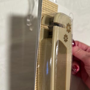 May include: A beige plastic paper punch with two gold flower accents. The punch is packaged in a clear plastic bag with a grid pattern.