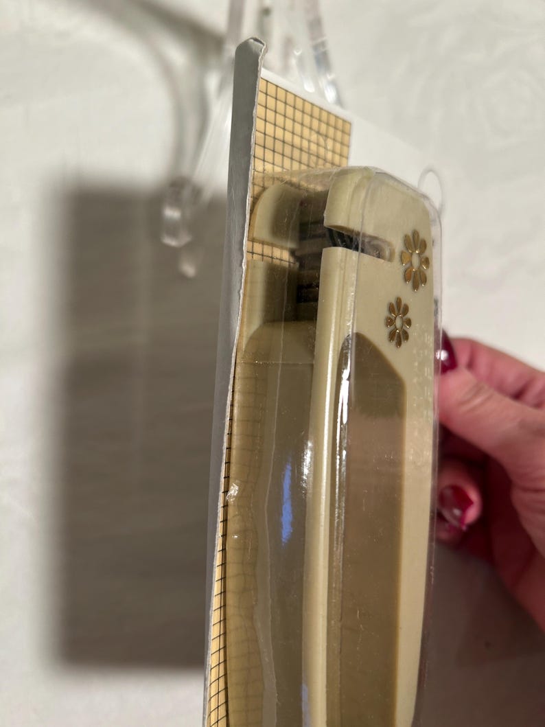 May include: A beige plastic paper punch with two gold flower accents. The punch is packaged in a clear plastic bag with a grid pattern.