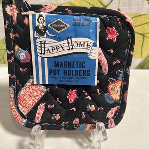 May include: A black quilted Happy Home magnetic pot holder with a colorful floral pattern. The pot holder has a loop for hanging and a blue and white label that reads "Happy Home Magnetic Pot Holders" and "Woolworths."