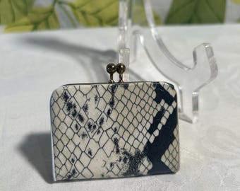 May include: A small, rectangular coin purse with a snakeskin pattern in black and cream. The purse has a gold-colored clasp closure. The background is a light-colored surface with a floral pattern.