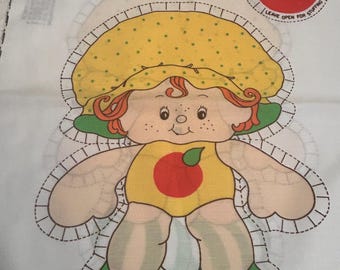 Op de afbeelding: Stoffpaneel met het personage "Apple Dumplin'" met een gele hoed, rood haar en een gele outfit met een rode appel. Het ontwerp bevat een rode appel met een groen blad en instructies om open te laten voor vulling.