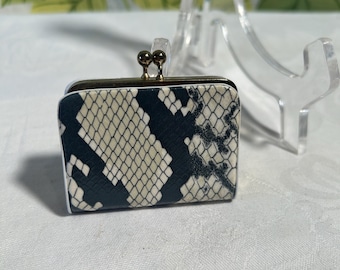 May include: A small, rectangular coin purse with a snakeskin pattern in black and cream. The purse has a gold-colored clasp with two small ball accents. The edges are trimmed in white.