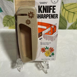 May include: A beige plastic knife sharpener with a floral design. The packaging says "SHARPIE" KNIFE SHARPENER and "Keep your kitchen and table knives sharp".