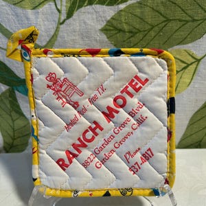 May include: A quilted, square pot holder with a yellow border and a white center. The center features red text that reads "RANCH MOTEL" and the address "8822 Garden Grove Blvd, Garden Grove, Calif." The pot holder has a loop for hanging.