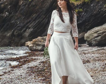 May include: A woman wearing a white lace crop top and a flowing white skirt stands on a rocky beach. The outfit is a bridal or formal wear ensemble. The background features a rocky cliffside and the ocean.