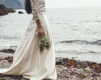 May include: A cream-colored wedding dress with a lace bodice and long skirt. The bride holds a small bouquet of greenery. The photo is taken on a beach with the ocean in the background.