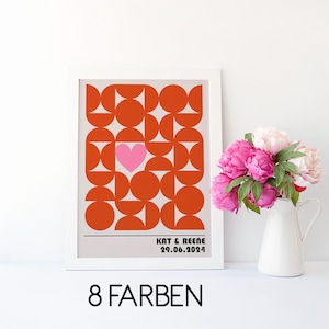 May include: A framed print with a geometric orange and pink design, featuring a pink heart. The print includes the names "KAT & REENE" and the date "29.06.2024". A white jug with pink flowers is next to the print.