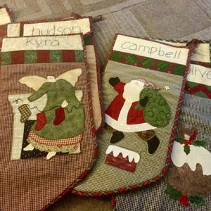 May include: A collection of quilted Christmas stockings featuring fabric appliqués of an angel, Santa Claus, and a holiday pudding. Each stocking has a white cuff with names like Kyra, Campbell, and Oliver written in black ink on the fabric.
