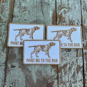 May include: Three rectangular embroidered patches with white fabric backgrounds and tan borders. Each patch features a gold-colored dog silhouette and the text "POINT ME TO THE BAR" stitched in gold thread across the bottom of the rectangular design.