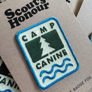 May include: A rectangular embroidered patch with the words "CAMP CANINE" and a tree design in green and white. The patch has a blue border and wavy lines at the bottom. The patch is on a brown card with the words "Scout's Honour" printed in black.