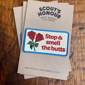 May include: A rectangular patch with a blue border and the text "Stop & smell the butts" with two red roses. The patch is on a tan card with the text "SCOUT'S HONOUR MERIT BADGES FOR DOGS".