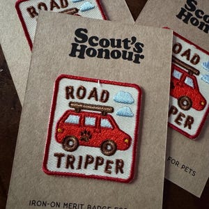 May include: Embroidered iron-on merit badges for pets. The badges feature a red car with the text "ROAD TRIPPER" and "ROAD" on top. The badges are on a brown card with the text "Scout's Honour".