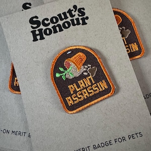 May include: A brown and orange iron-on patch with a cartoon illustration of a plant growing out of a pot with the text "Plant Assassin" on it. The patch is on a brown card with the text "Scout's Honour" and "Iron-on Merit Badge for Pets".