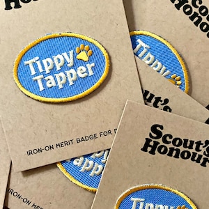 May include: Three iron-on merit badges for dogs. The badges are blue with a yellow border and feature a paw print and the text "Tippy Tapper" in white. The badges are on brown card stock with the text "Scout's Honour" and "IRON-ON MERIT BADGE FOR DOGS" printed on them.