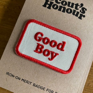 May include: White iron-on merit badge for dogs with red embroidered text that says "Good Boy" and a red border. The badge is on a brown card with the text "Scout's Honour" and "IRON-ON MERIT BADGE FOR DOGS".