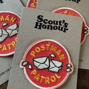 May include: Red and yellow embroidered iron-on patch with the text "Postman Patrol" and two white envelopes. The patch is on a brown card with the text "Scout's Honour" and "Iron-on Merit Badge for Dogs".