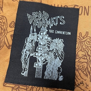 May include: Black and white patch with the text "Dead Formats VHS Convention" and an illustration of two figures with long hair, one standing on a pole with VHS tapes stacked on it, and the other holding a VHS tape.