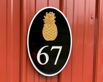 Könnte beinhalten: Ein ovales Schild in Schwarz und Weiß mit einem goldenen Ananas-Design und der Nummer 67. Das Schild ist an einer roten Metalloberfläche befestigt.