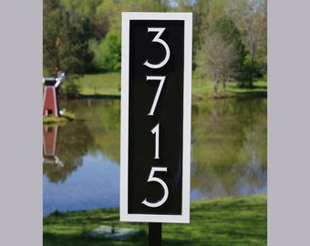May include: Black and white house number sign with the numbers 3715 displayed in a vertical column. The sign is mounted on a black post and is in front of a pond and green grass.