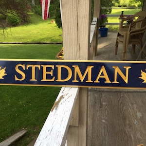 May include: A blue wooden sign with gold lettering and maple leaf accents. The sign reads "STEDMAN".