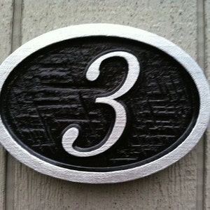 May include: A silver-rimmed, black circular sign with the number "3" in white cursive lettering.
