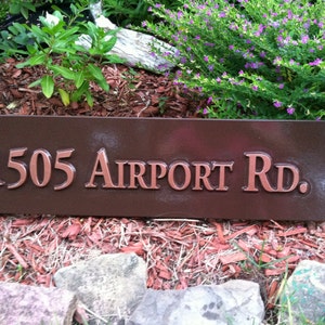 May include: A brown wooden sign with the address "1505 Airport Rd." in copper letters.