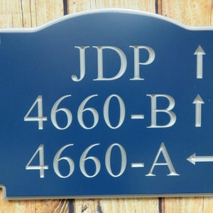 May include: A navy blue address sign with a decorative edge. The sign features the engraved text "JDP", "4660-B", and "4660-A", along with directional arrows. The background is a wooden surface.