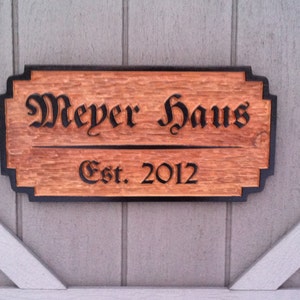 May include: A wooden sign with black lettering that reads "Meyer Haus Est. 2012". The sign has a dark brown border and is mounted on a light gray wall.