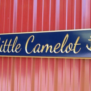 May include: A blue wooden sign with gold lettering and anchors on either side. The sign reads "Little Camelot".