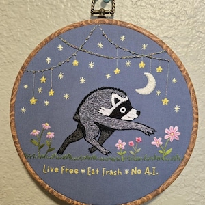 May include: A circular embroidery hoop featuring a grey and black raccoon running across a field of pink flowers under a night sky with yellow stars and a white crescent moon. The text "Live Free * Eat Trash * No A.I." is stitched in yellow.