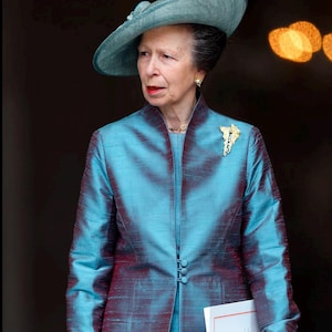 Puede incluir: Una mujer con un traje de seda color turquesa y sombrero a juego, con una broche dorada. Sostiene un bolso de mano crema y un documento blanco. El conjunto es formal.