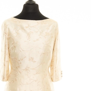 May include: A cream-colored dress with a floral pattern. The dress has a scoop neck and three-quarter sleeves. The dress is on a mannequin.