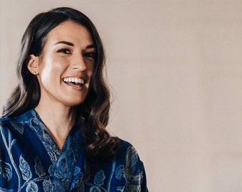 May include: A woman with long dark hair smiles while wearing a blue and white floral patterned robe.
