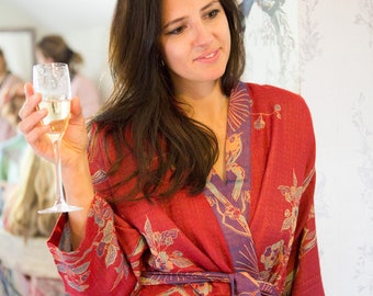 May include: A woman wearing a red and purple floral robe holds a glass of champagne. The robe has a long, flowing design and a tie belt.