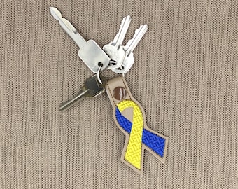 May include: A beige leather keychain featuring an embroidered awareness ribbon in bright yellow and blue with a textured basketweave pattern. The keychain is attached to a silver metal split ring holding three silver metal keys of varying sizes.